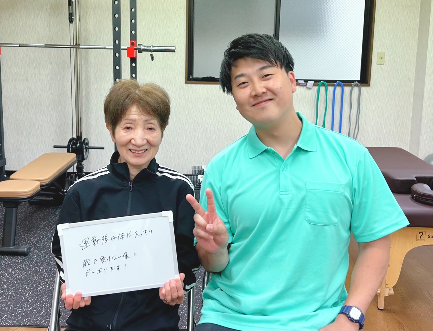 年配の女性とトレーナーが、手書きのメモが書かれたホワイトボードを手に、カメラに向かって微笑んでいます。背景にはジムの器具が映っています。