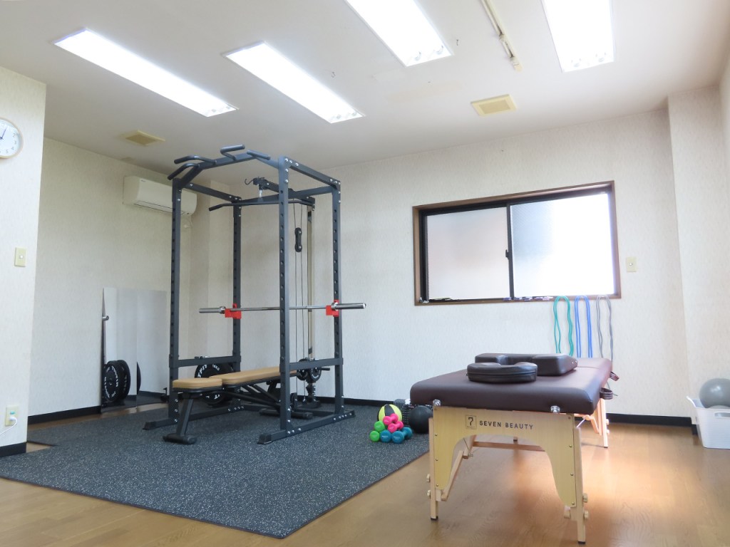 室内に設置されたジムの器具と施術台の画像。トレーニング器具、バーベル、カラフルなエクササイズボールやチューブが見える。