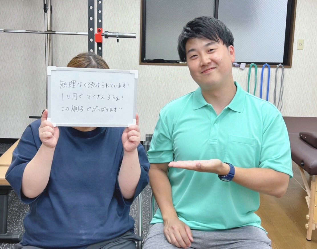 女性が白いボードを持っている写真。ボードには、1ヶ月で体重が3kg減少したことを報告する内容が手書きで書かれている。隣には男性のトレーナーが座っていて、笑顔を見せている。