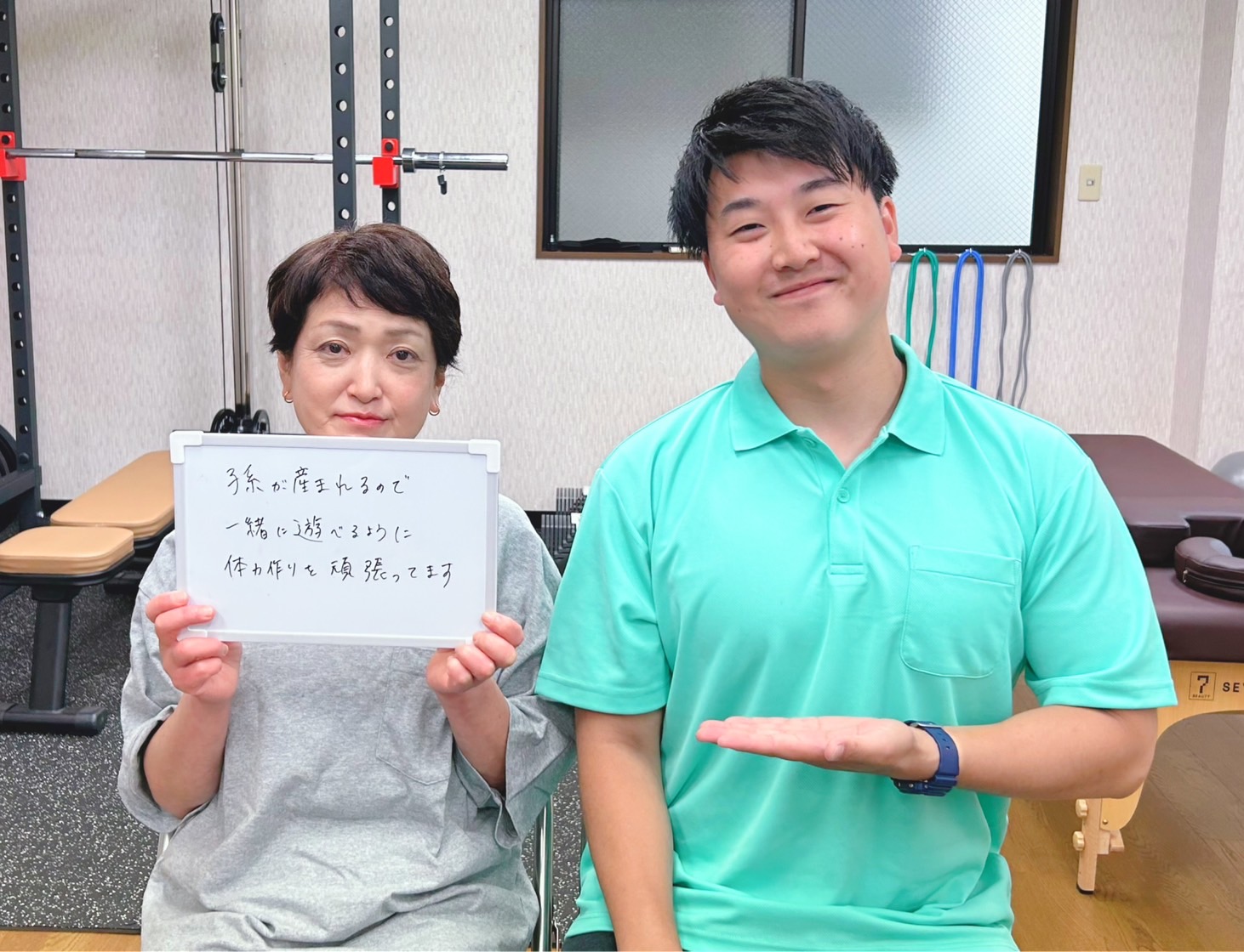 女性が白いボードを持ち、トレーナーと笑顔でポーズをとるジムの内部での写真。背景にはトレーニング器具が見える。