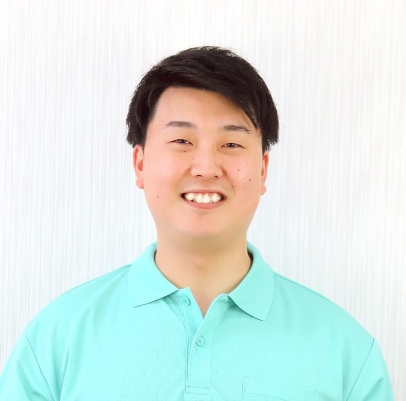 明るい色の背景の前に立って、水色のポロシャツを着た笑顔の男性。