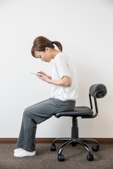 女性が椅子に座り、スマートフォンを見ている姿勢。